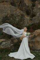 le la mariée est permanent près une Roche haute dans le montagnes, suivant à une grand pierre. mariage robe. le vent larmes le voile photo