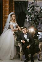 portrait de positif Jeune jeune marié dans noir costume avec blanc chemise avec la mariée souriant et séance sur confortable foncé vert canapé dans moderne Hôtel pièce sur mariage journée photo