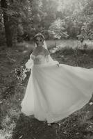 une magnifique la mariée dans une blanc robe avec une longue voile est encerclant avec une bouquet dans le parc. Jeune femme, art photo, mariage, mariée, ouvert épaules. lumière. noir et blanc photo