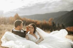 portrait de une élégant jeune marié avec une la mariée sur une Contexte de l'automne sec herbe. le concept de une rural mariage dans le montagnes, content bohémien jeunes mariés. le la mariée et jeune marié sont mensonge sur le herbe photo