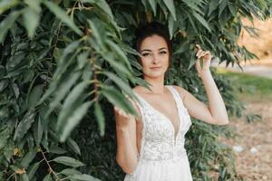 mariage photo sur une vert Contexte. le la mariée dans une blanc robe des stands près vert des buissons, détient une feuille et regards dans le lentille. magnifique se maquiller. portrait de le la mariée