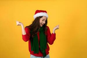magnifique content asiatique femme dans rouge Noël chandail et chapeau. elle est excité et serre tous les deux les poings sur une Jaune studio Contexte. photo