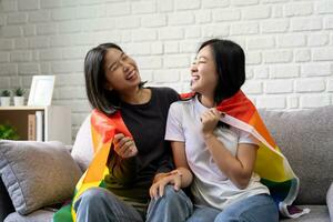 content lesbiennes, deux Jeune asiatique femmes en portant arc en ciel lgbt drapeau avec fierté ensemble. elles ou ils étaient séance sur le canapé à maison. photo
