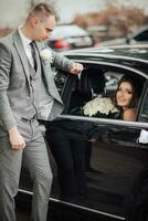 mariage photo. le la mariée avec une bouquet de des roses est assis souriant dans une noir voiture, le jeune marié des stands penché sur le voiture et regards à le la mariée. magnifique maquillage et cheveux. une élégant homme photo