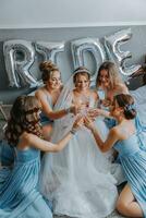 Jeune demoiselles d'honneur dans bleu soie Robes boisson Champagne dans le de la mariée chambre. magnifique femmes célébrer bachelorette fête séance sur lit et avec Champagne. photo