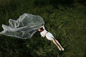 Matin de le la mariée dans la nature. le la mariée est dans une pansement robe et une longue voile, mensonge sur le vert herbe et profiter le du soleil chaleur. large angle photo. photo