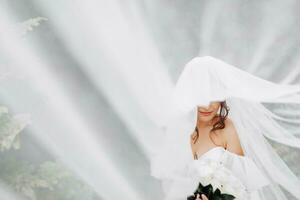 frisé brunette la mariée dans une blanc robe, couvert avec une voile, pose pour le caméra avec une bouquet de des roses. portrait de le la mariée. magnifique maquillage et cheveux. mariage dans la nature photo