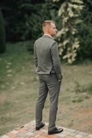 une homme dans une gris costume pose dans la nature avec le sien mains dans le sien les poches, le sien épaules tourné à le caméra. arrière voir. une élégant montre. Pour des hommes style. mode. affaires photo