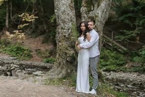 une fille avec longue noir frisé cheveux, permanent dans une blanc soie robe dans une printemps forêt contre une grand arbre, détient une fougère branche dans sa mains. derrière sa est sa mari photo