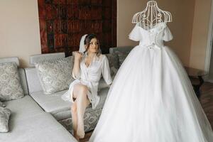 une brunette la mariée dans une pansement robe est assis sur une gris canapé, en portant sa noir boucles derrière une mannequin avec une mariage robe, contre le Contexte de peint des murs. mariage photo. magnifique la mariée photo