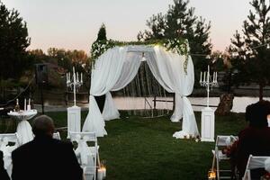 mariage la cérémonie sur le rive de le lac. La peinture la cérémonie dans le soir. romantique peinture. le présidium est décoré avec blanc fleurs. blanc chaises photo