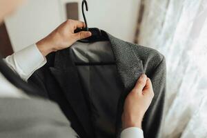 une homme dans une blanc chemise détient le sien gris veste, contre le Contexte de une la fenêtre. de face voir. une élégant montre. Pour des hommes style. mode. affaires photo