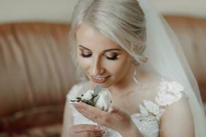 magnifique blond la mariée portrait, en portant lacet dans main mariage maquillage coiffure, magnifique Jeune femme dans blanc robe à maison. séries photo