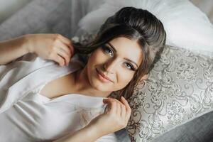 brunette la mariée dans une pansement robe posant pour le caméra tandis que mensonge sur une gris canapé. portrait photo. élégant coiffure. agréable se maquiller. magnifique mains. Matin de le la mariée. photo