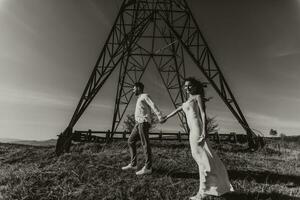 élégant modèle couple dans le montagnes dans été. une Jeune garçon et fille dans une blanc soie robe sont permanent près grand les structures de Puissance lignes. noir et blanc contraste photo