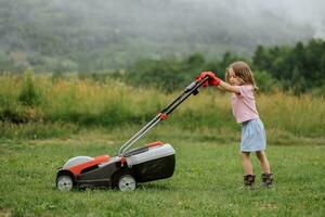 une enfant dans bottes dans le forme de une Jeu tond herbe avec une tondeuse à gazon dans le Cour contre le Contexte de montagnes et brouillard, le concept de jardin outils photo