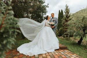 plein longueur côté vue de un magnifique sensuel Jeune brunette la mariée dans longue blanc mariage robe et voile permanent dans parc en portant bouquet ensemble avec jeune marié en plein air sur Naturel arrière-plan, horizontal photo