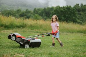 une enfant dans bottes dans le forme de une Jeu tond herbe avec une tondeuse à gazon dans le Cour contre le Contexte de montagnes et brouillard, le concept de jardin outils photo