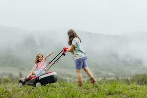 une femme dans bottes avec sa enfant dans le forme de une Jeu tond le herbe avec une tondeuse à gazon dans le jardin contre le Contexte de montagnes et brouillard, jardin outils concept photo