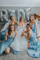 Jeune demoiselles d'honneur dans bleu soie Robes boisson Champagne dans le de la mariée chambre. magnifique femmes célébrer bachelorette fête séance sur lit et avec Champagne. photo