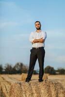 mariage portrait photo. une élégant jeune marié dans une blanc chemise pose sur une foins balle contre une bleu ciel Contexte. barbu homme. style. photo