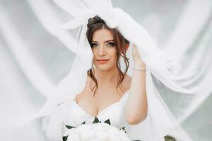 frisé brunette la mariée dans une blanc robe, couvert avec une voile, pose pour le caméra avec une bouquet de des roses. portrait de le la mariée. magnifique maquillage et cheveux. mariage dans la nature photo