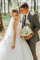 à la mode jeune marié et mignonne blond la mariée dans une blanc robe avec une couronne, une bouquet sont étreindre, en riant dans le parc, jardin, forêt en plein air. mariage la photographie, portrait de souriant jeunes mariés. photo