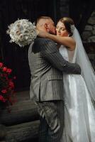 une romantique la mariée dans une blanc robe avec une bouquet embrasse le jeune marié près le en bois porte de le église entrée photo