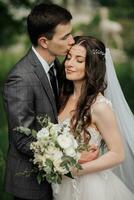 content Jeune couple. mariage portrait. le jeune marié baisers le la mariée sur le temple. Jeune gens sont tendrement étreindre tandis que à la recherche à le caméra. mariage bouquet. printemps mariage photo