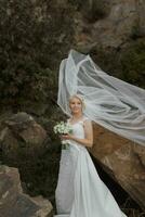 le la mariée est séance sur une Roche haute dans le montagnes suivant à une grand pierre. mariage robe. le vent larmes le voile photo