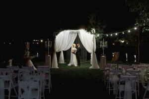 mariage la cérémonie sur le rive de le lac. La peinture la cérémonie dans le soir. romantique peinture. le présidium est décoré avec blanc fleurs. blanc chaises. le la mariée et jeune marié doucement tenir mains photo