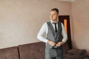 une homme dans une blanc chemise et une gris gilet pose par le fenêtre et regards à le caméra, ajuste le sien gilet. de face voir. une élégant montre. Pour des hommes style. mode. affaires photo