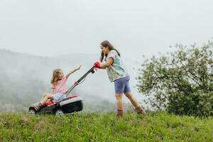 une femme dans bottes avec sa enfant dans le forme de une Jeu tond le herbe avec une tondeuse à gazon dans le jardin contre le Contexte de montagnes et brouillard, jardin outils concept photo