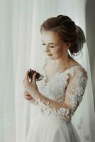 préparation pour le mariage. magnifique Jeune la mariée dans blanc mariage robe à l'intérieur. luxe modèle regards à mariage chaussures, à Accueil dans studio pièce avec gros la fenêtre. le fille spectacles photo