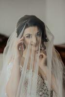 portrait. une brunette la mariée dans une blanc robe, enveloppé dans une voile, regards vers le bas et touche sa affronter, pose. magnifique maquillage et cheveux. volumineux voile. mariage photo. magnifique la mariée photo