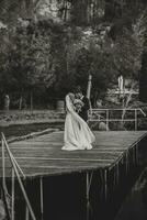 jeune marié dans une noir costume et blanc chemise dansant sur une jetée avec une la mariée dans une blanc mariage robe, près une Lac et rochers. noir et blanc photo