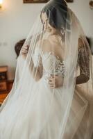 portrait. une brunette la mariée dans une blanc robe, enveloppé dans une voile, regards vers le bas et touche sa affronter, pose. magnifique maquillage et cheveux. volumineux voile. mariage photo. magnifique la mariée photo