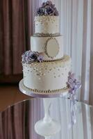 blanc à trois niveaux mariage gâteau décoré avec violet fleurs et perles, permanent sur une miroir rester. desserts mariage. photo