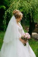 large portrait de le la mariée. le la mariée est permanent dans une blanc robe et voile avec une bouquet et à la recherche à lui, contre le Contexte de luxuriant vert des arbres. une magnifique robe. jolie femme. photo