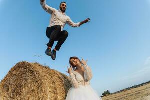 mariage photo. une élégant jeune marié dans une blanc chemise sauts vers le bas de une foins balle, le la mariée est permanent dessous. barbu homme. roux la mariée. style. émotions photo