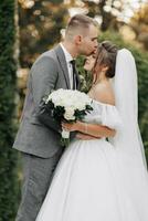 portrait de le la mariée et jeune marié dans la nature. le jeune marié et le brunette la mariée dans une blanc longue robe sont debout, embrasser contre le Contexte de conifères. élégant jeune marié. magnifique cheveux et maquillage photo