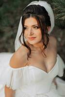 portrait de le la mariée dans la nature. une brunette la mariée dans une blanc volumineux robe est séance, posant près une conifère arbre, en portant une bouquet de blanc des roses. magnifique cheveux et se maquiller. chaud lumière photo