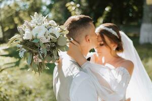 portrait de une magnifique couple dans l'amour sur votre mariage journée. une marcher dans le parc dans le ensoleillement et verdure. incroyable baisers et câlins de le la mariée et jeune marié avec une bouquet photo