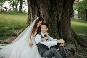 une magnifique la mariée avec longue frisé cheveux dans une élégant robe câlins le marié, sourires, à la recherche dans le lentille en dessous de une gros arbre. portrait de le la mariée et jeune marié. printemps mariage. Naturel maquillage photo