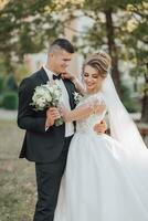 mariage portrait. le jeune marié dans une noir costume et le blond la mariée sont debout, étreindre, posant en portant une bouquet en dessous de une arbre. photo session dans la nature. magnifique cheveux et maquillage