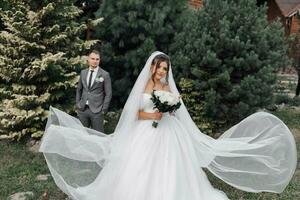 une aux cheveux bouclés brunette la mariée dans une blanc robe et voile, écoulement magnifiquement dans le air, des stands dans de face de sa élégant jeune marié. portrait de le la mariée et jeune marié. mariage dans la nature photo