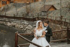 une élégant la mariée dans une luxuriant robe et à la mode coiffure des stands sur une jetée avec une jeune marié dans une parc près en bois Maisons, fermer coup photo