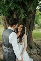 une magnifique la mariée avec longue frisé cheveux dans un élégant robe regards à le jeune marié avec aimant yeux, sourit. portrait de le la mariée et jeune marié. printemps mariage. Naturel maquillage photo