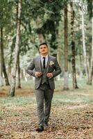 une élégant jeune marié des promenades vers l'avant dans le forêt boutonnage le sien veste entre grand des arbres. une à la mode marié, un adulte homme photo