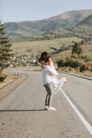 une élégant jeune marié dans une blanc chemise et une mignonne brunette la mariée dans une blanc robe sont encerclant sur un asphalte route contre le Contexte de une forêt et montagnes. mariage portrait de jeunes mariés. photo